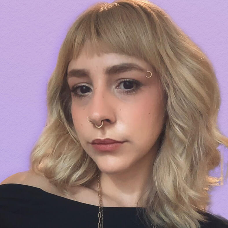A foto foi tirada de baixo, temos uma mulher em primeiro plano olhando diretamente pra câmera, o cabelo é preto na altura dos ombros com uma mecha loira. Ela tem franja, usa maquiagem leve e uma blusa preta. Tem piercing no septo, aba nasal e sobrancelha.